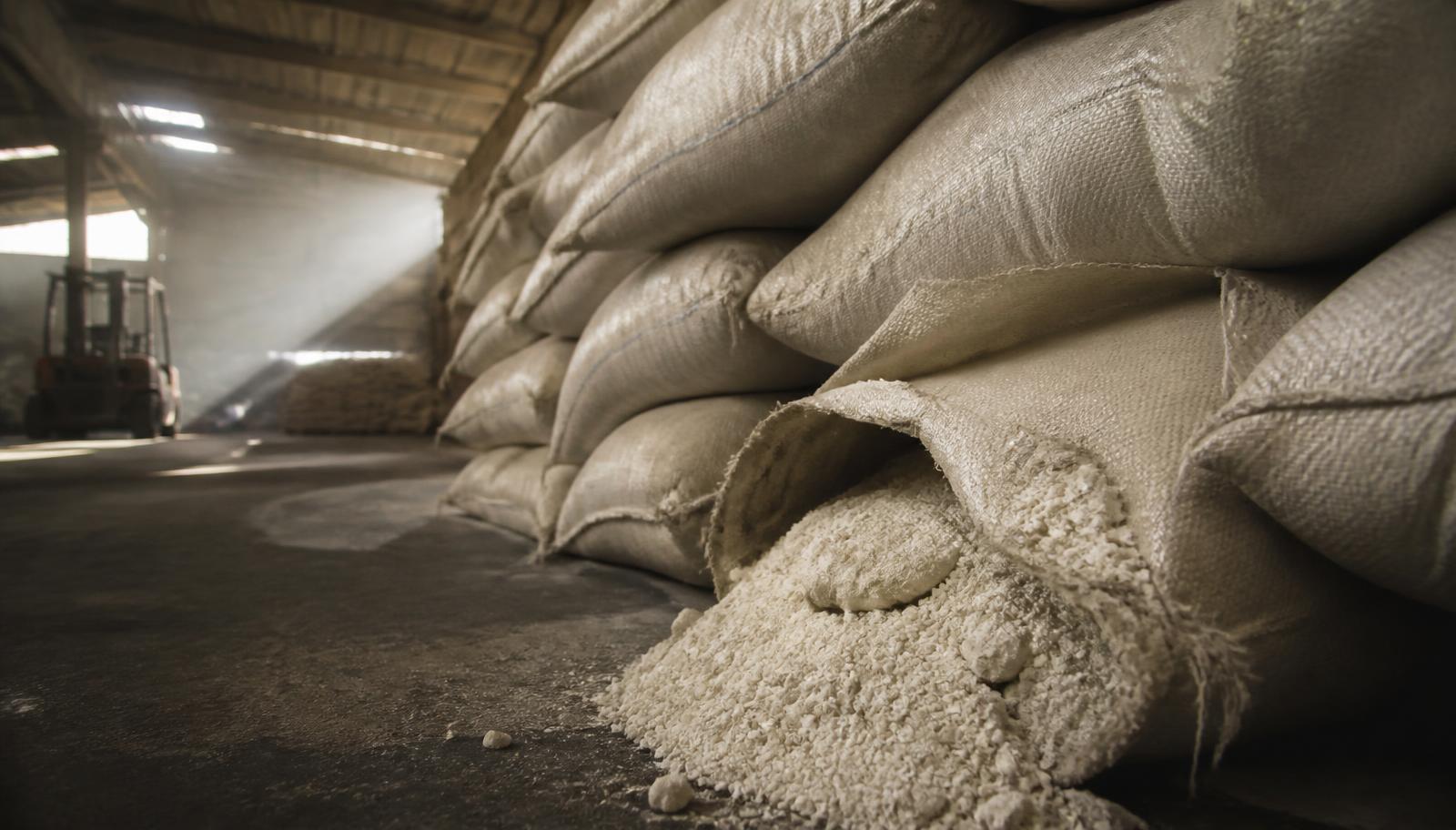 Stacked unmarked woven sacks of fertiliser in a port warehouse with one split bag showing caked hardened granules