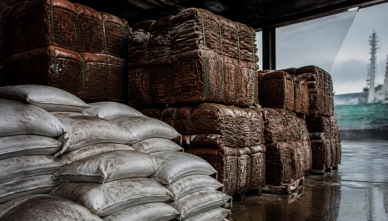 Stacked bales of natural rubber and unbranded sacks of raw material in a humid port warehouse