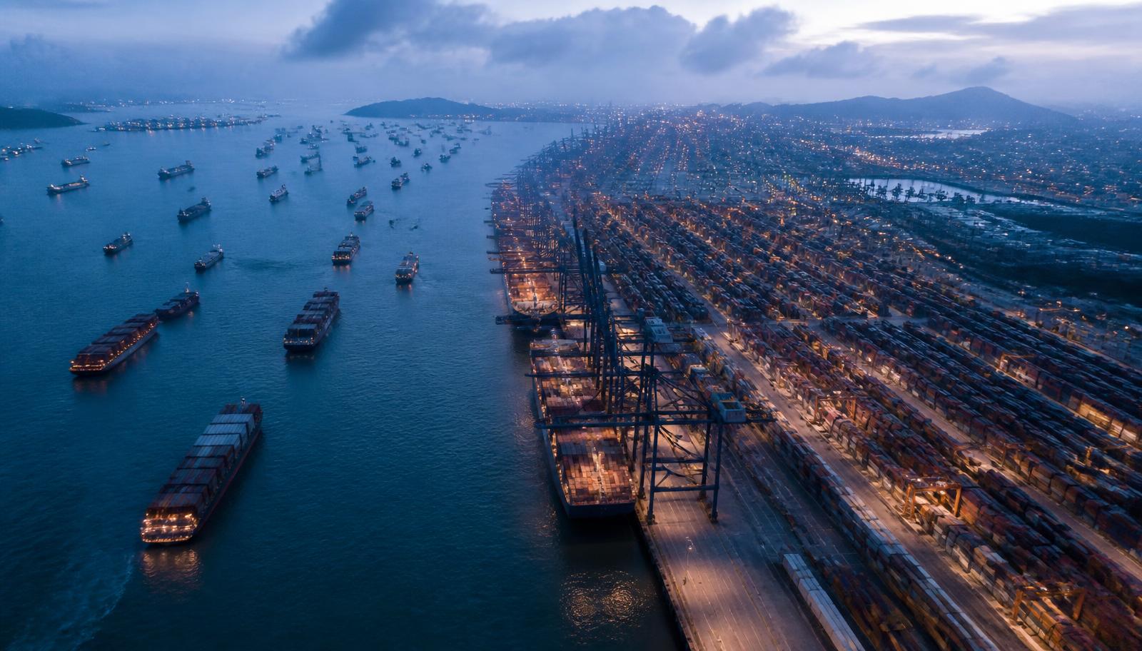 Aerial view of a large container terminal at blue hour with unbranded container vessels at anchor offshore